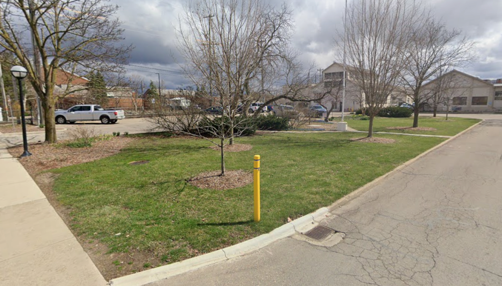 A grassy lawn area called the Physical Properties Building Lawn is shown beside a paved road. Several young trees are planted throughout the lawn, each surrounded by mulch. On the left, a sidewalk runs parallel to the street and is bordered by a lamp post. A bright yellow bollard stands near the curb in the foreground. In the background, there are parked vehicles, shrubs, and multiple buildings.