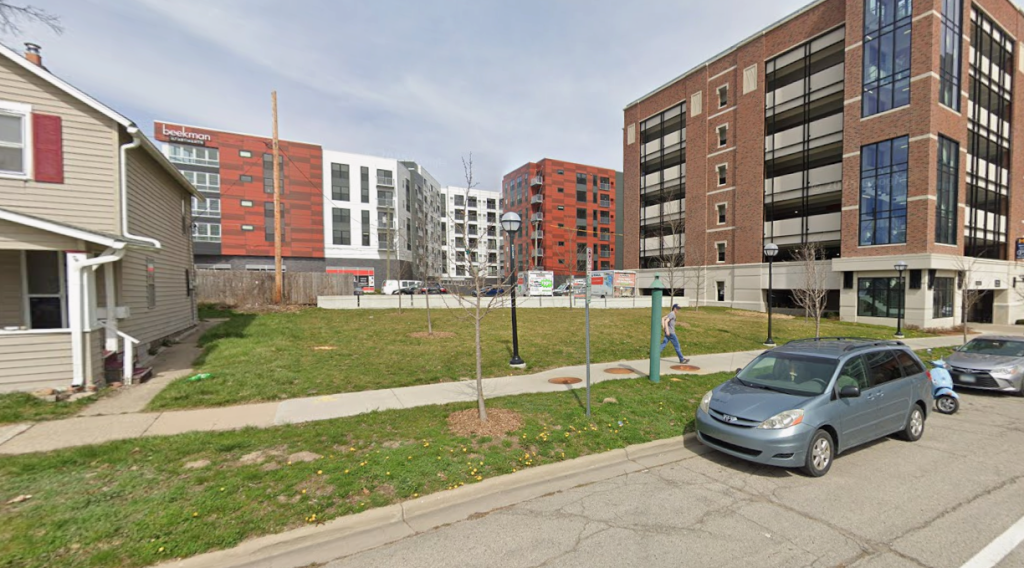 A grassy area known as the Wall Street West Lawn sits alongside a street. Several young trees grow along its sidewalk, which runs parallel to the curb. On the left, part of a beige house is visible. The large brown brick building on the right is Parking Structure M99, featuring tall windows.